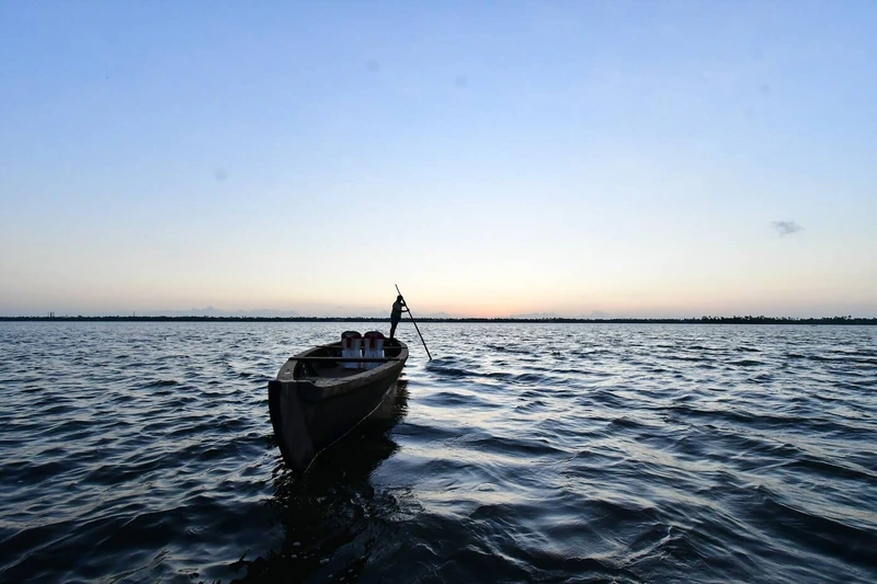 A traditional wooden boat poled across the calm backwaters at dusk, silhouetted against a pale sky