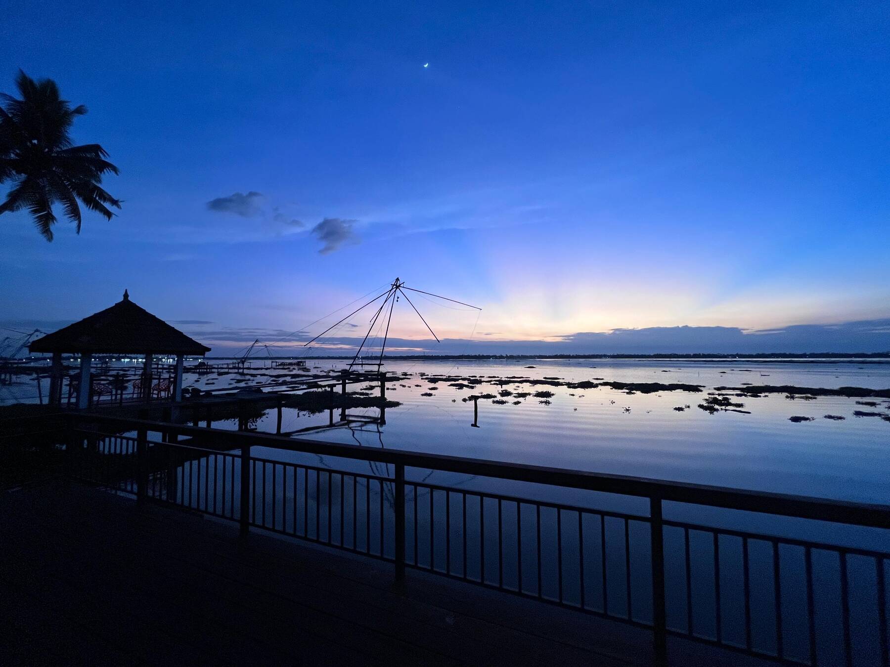 A Chinese fishing net and waterside pavilion silhouetted against the blue hour sky over the Kumbalangi backwaters